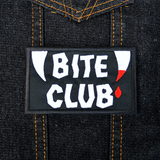 Rectangular black embroidered patch with the words bite club sewn in white thread with a pair of fangs on either side, one of which is dripping blood.