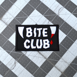 Rectangular black embroidered patch with the words bite club sewn in white thread with a pair of fangs on either side, one of which is dripping blood.