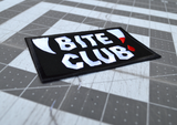 Close up detail of a rectangular black embroidered patch with the words bite club sewn in white thread with a pair of fangs on either side, one of which is dripping blood.