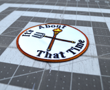 Close up detail of a circular embroidered patch with a brown border, a torch and pitchfork crossed over each other in the middle, surrounded by the phrase, it's about that time.