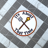 Circular embroidered patch with a brown border, a torch and pitchfork crossed over each other in the middle, surrounded by the phrase, it's about that time.