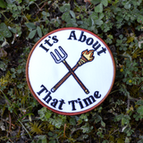Circular embroidered patch with a brown border, a torch and pitchfork crossed over each other in the middle, surrounded by the phrase, it's about that time.