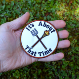 Circular embroidered patch with a brown border, a torch and pitchfork crossed over each other in the middle, surrounded by the phrase, it's about that time. Shown in the palm of a hand for size reference.