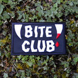 Rectangular black embroidered patch with the words bite club sewn in white thread with a pair of fangs on either side, one of which is dripping blood.