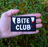 Rectangular black embroidered patch with the words bite club sewn in white thread with a pair of fangs on either side, one of which is dripping blood. Shown in the palm of a hand for size reference.