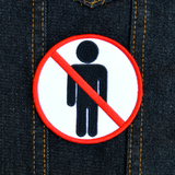 Circular white embroidered patch with red border and a red line crossing out a black silhouette of a person, resembling a public sign. Shown on a denim jacket.