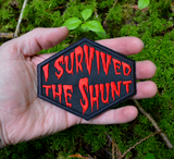 Red on black embroidered patch that says I survived the shunt, in reference to the horror movie, Society. Held in the palm of a hand for size reference.