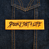 Rectangular orange embroidered patch with the text, spooky shit 4 life, stitched in black thread. Shown on a denim vest.