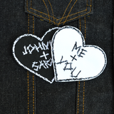 Two heart-shaped embroidered patches with personalized names for couples. One is white stitching on black fabric, one is black stitching on white fabric.