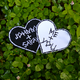 Two heart-shaped embroidered patches with personalized names for couples. One is white stitching on black fabric, one is black stitching on white fabric.