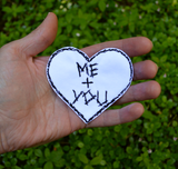 A custom black and white personalized heart-shaped embroidered patch with 'ME + YOU' text held in a hand against a green leafy background.