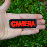 Black rectangular embroidered patch with the name Gamera in red thread, based on the classic Japanese Kaiju monster movie. Shown in the palm of a hand for size reference.