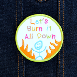 Circular patch with text 'Let's Burn it All Down' with a happy stick figure and flames on a white background and green border. Shown on a denim vest.