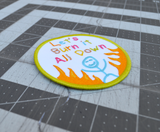 Close up detail of a circular patch with text 'Let's Burn it All Down' with a happy stick figure and flames on a white background and green border.
