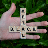An embroidered patch that says black metal spelled out in Scrabble letters in the shape of an inverted cross. Shown in the palm of a hand for size reference.