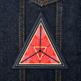 A triangular embroidered patch with a black border, textured red background and geometric designs in silver and gold metallic thread.