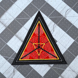 A triangular embroidered patch with a black border, textured red background and geometric designs in silver and gold metallic thread.
