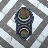 An oblong embroidered patch with abstract geometric arcs and circles in grey, black, and multi-color thread.