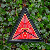 A triangular embroidered patch with a black border, textured red background and geometric designs in silver and gold metallic thread.