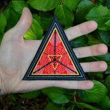 A triangular embroidered patch with a black border, textured red background and geometric designs in silver and gold metallic thread. Held in the palm of a hand for size reference.