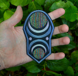 An oblong embroidered patch with abstract geometric arcs and circles in grey, black, and multi-color thread. Shown in the palm of a hand for size reference.