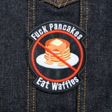 A round-shaped embroidered patch with a red circle and slash over a plate of pancakes surrounded by the words, fuck pancakes, eat waffles.