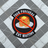 A round-shaped embroidered patch with a red circle and slash over a plate of pancakes surrounded by the words, fuck pancakes, eat waffles.