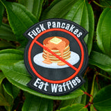 A round-shaped embroidered patch with a red circle and slash over a plate of pancakes surrounded by the words, fuck pancakes, eat waffles.