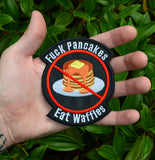 A round-shaped embroidered patch with a red circle and slash over a plate of pancakes surrounded by the words, fuck pancakes, eat waffles. Shown in the palm of a hand for size reference.
