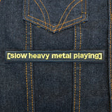 A rectangular black embroidered patch with the words slow heavy metal playing, stitched in yellow thread to resemble a movie subtitle.