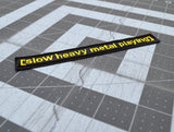 Close up detail of a rectangular black embroidered patch with the words slow heavy metal playing, stitched in yellow thread to resemble a movie subtitle.