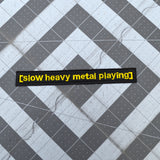A rectangular black embroidered patch with the words slow heavy metal playing, stitched in yellow thread to resemble a movie subtitle.