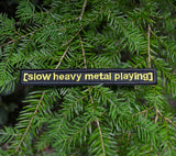 A rectangular black embroidered patch with the words slow heavy metal playing, stitched in yellow thread to resemble a movie subtitle.