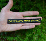 A rectangular black embroidered patch with the words slow heavy metal playing, stitched in yellow thread to resemble a movie subtitle. Held in the palm of a hand for size reference.