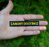 Rectangular black embroidered patch with yellow lettering that says angry dooting, resembling a movie subtitle. Shown in the palm of a hand for size reference.