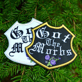 A pair of emblem-shaped embroidered patches with purple flowers and the words Got the Morbs in old english lettering. One is white with a silver border, the other black with a gold border.