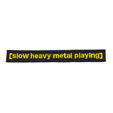 A rectangular black embroidered patch with the words slow heavy metal playing, stitched in yellow thread to resemble a movie subtitle.