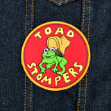 Circular patch on red fabric and gold border featuring a toad being stepped on by a boot and the words 'toad stompers' stitched in yellow thread, as a reproduction from the sci-fi movie Hell Comes to Frogtown.