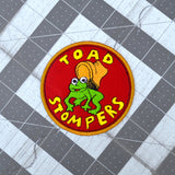 Circular patch on red fabric and gold border featuring a toad being stepped on by a boot and the words 'toad stompers' stitched in yellow thread, as a reproduction from the sci-fi movie Hell Comes to Frogtown.