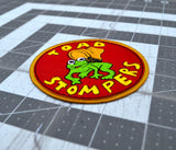 Close up detail of a circular patch on red fabric and gold border featuring a toad being stepped on by a boot and the words 'toad stompers' stitched in yellow thread, as a reproduction from the sci-fi movie Hell Comes to Frogtown.
