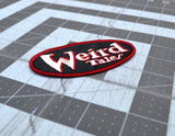 Close up detail of an oval shaped embroidered patch on black fabric, a red border and the logo for the classic strange fiction magazine, Weird Tales stitched in white and red.