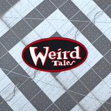 Oval shaped embroidered patch on black fabric, a red border and the logo for the classic strange fiction magazine, Weird Tales stitched in white and red.