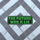 A rectangular embroidered patch on black fabric and a green border, with the words 'the future was a lie' in green block letters.