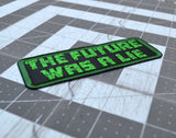 Close up detail of a rectangular embroidered patch on black fabric and a green border, with the words 'the future was a lie' in green block letters.
