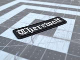 Close up detail of a black rectangular embroidered patch with the word 'therewolf' stitched with white thread in Old English lettering.