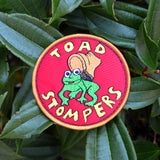 Circular patch on red fabric and gold border featuring a toad being stepped on by a boot and the words 'toad stompers' stitched in yellow thread, as a reproduction from the sci-fi movie Hell Comes to Frogtown.