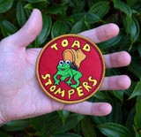 Circular patch on red fabric and gold border featuring a toad being stepped on by a boot and the words 'toad stompers' stitched in yellow thread, as a reproduction from the sci-fi movie Hell Comes to Frogtown. Shown in the palm of a hand for size reference.