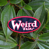Oval shaped embroidered patch on black fabric, a red border and the logo for the classic strange fiction magazine, Weird Tales stitched in white and red.