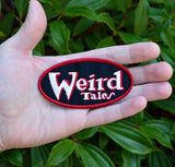 Oval shaped embroidered patch on black fabric, a red border and the logo for the classic strange fiction magazine, Weird Tales stitched in white and red. Shown in the palm of a hand for size reference.