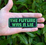 A rectangular embroidered patch on black fabric and a green border, with the words 'the future was a lie' in green block letters. Shown in the palm of a hand for size reference.